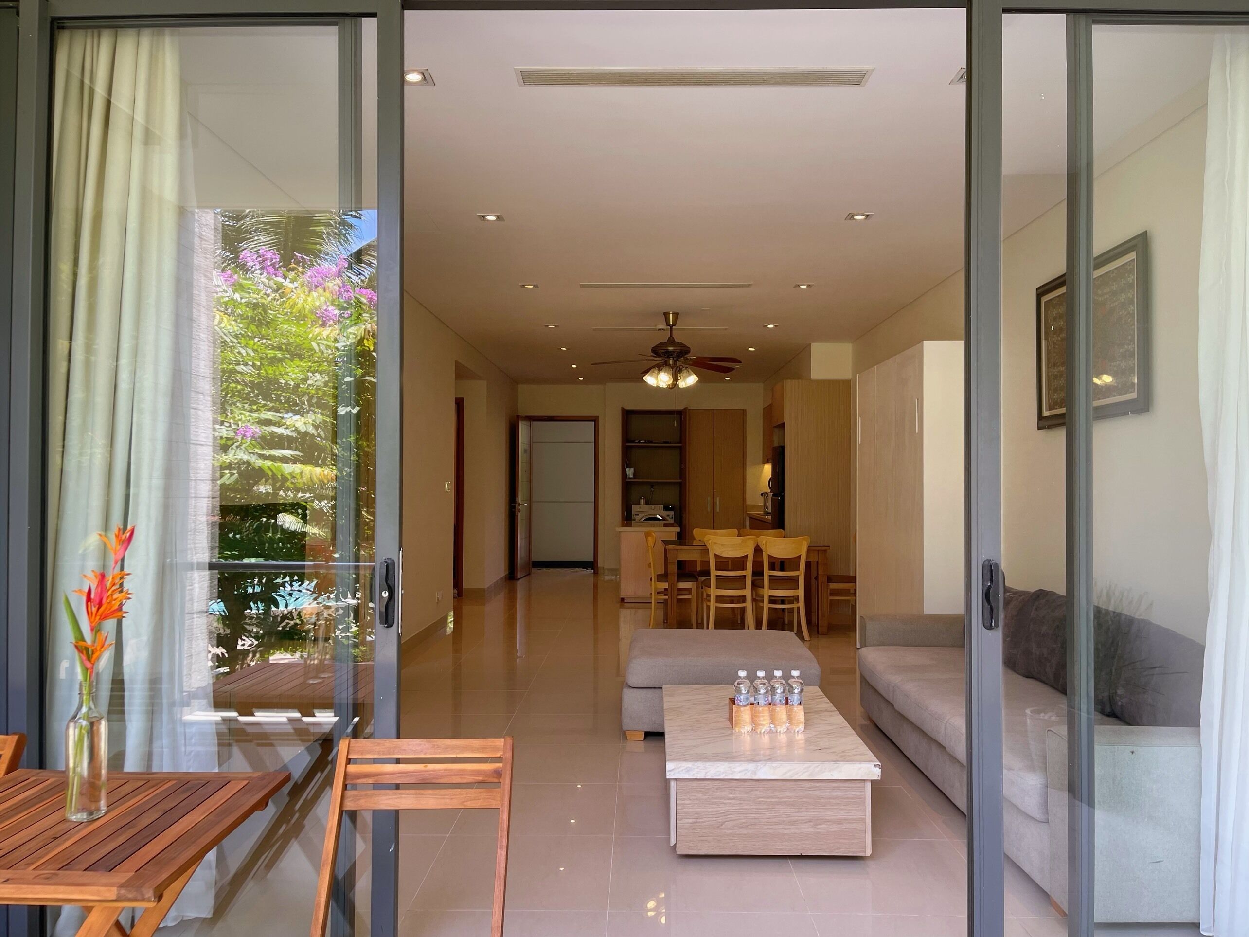 A modern living room with a glass door leading to a balcony, a dining area, and a kitchen in the background.