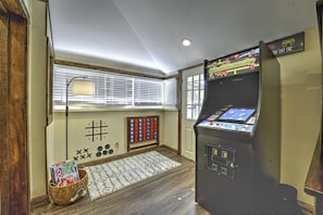 Ein Zimmer mit Flippermaschine, einem Fenster mit Jalousien, einer Lampe, einem gemusterten Teppich und einem Korb mit Zeitschriften.