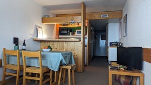 A compact kitchen with wooden cabinets, a microwave, and a small refrigerator. There is a dining area with a table and chairs, and a television on a wooden stand.
