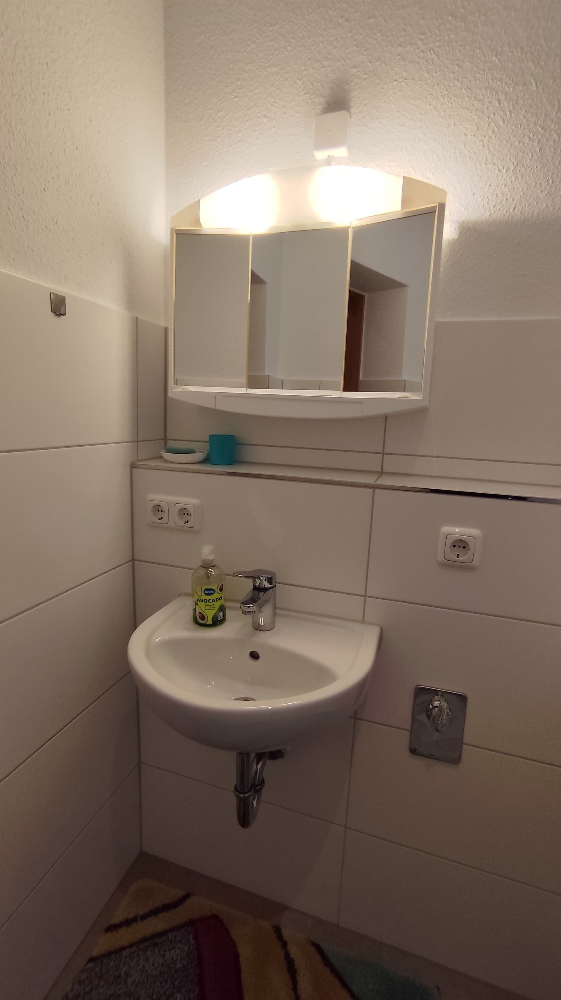 A bathroom with a white sink, a mirror, and a light above it. There are two electrical outlets on the wall and a small shelf with a bowl and a cup.