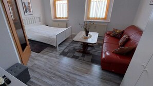 A compact living space with a bed, a red sofa, a small table, and a view of the outside through a window. - Ferienwohnung Modern (Burgstädt)