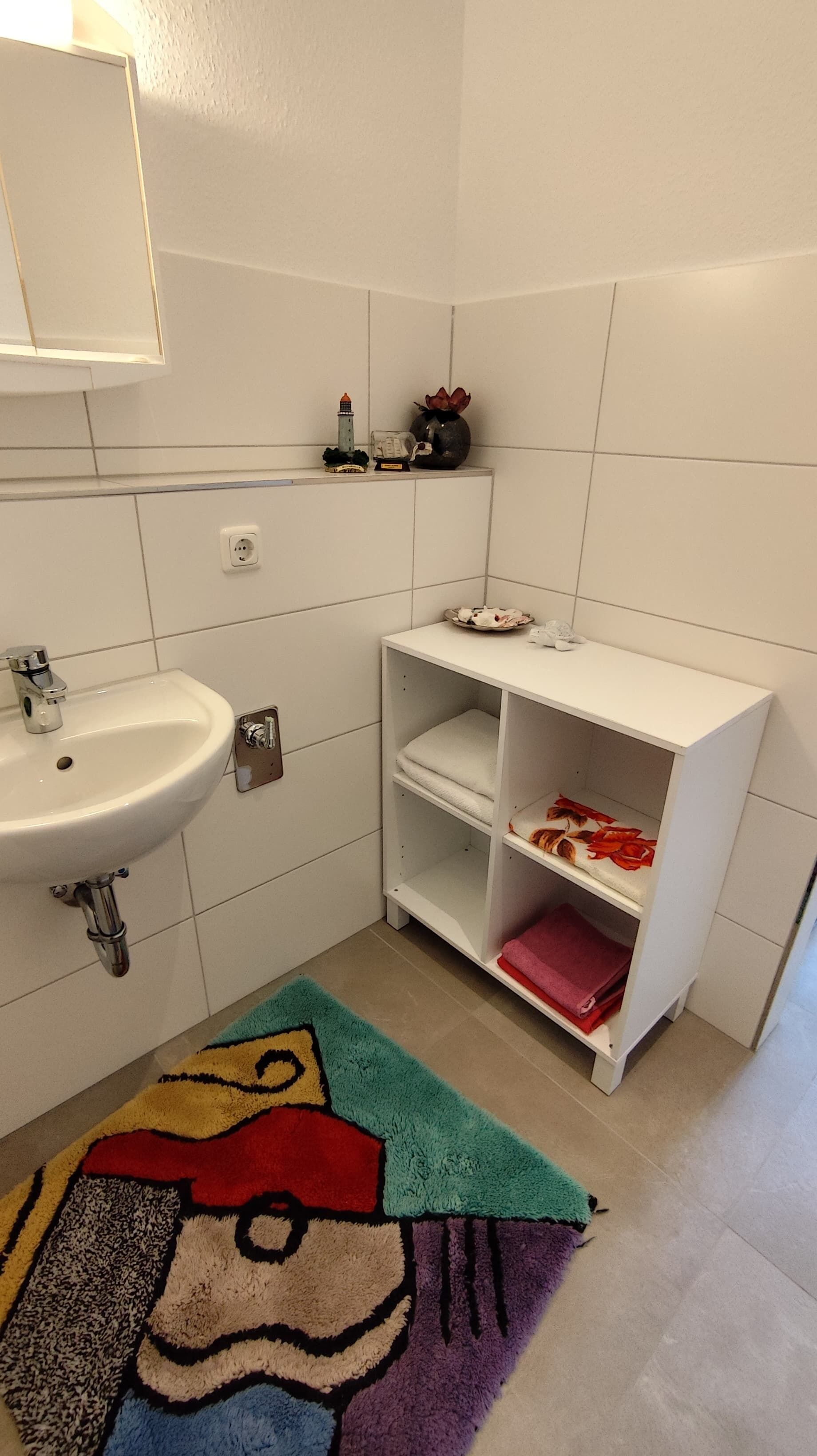 Un baño con un lavamanos blanco, un espejo encima, una repisa con adornos y una alfombra colorida en el suelo.
