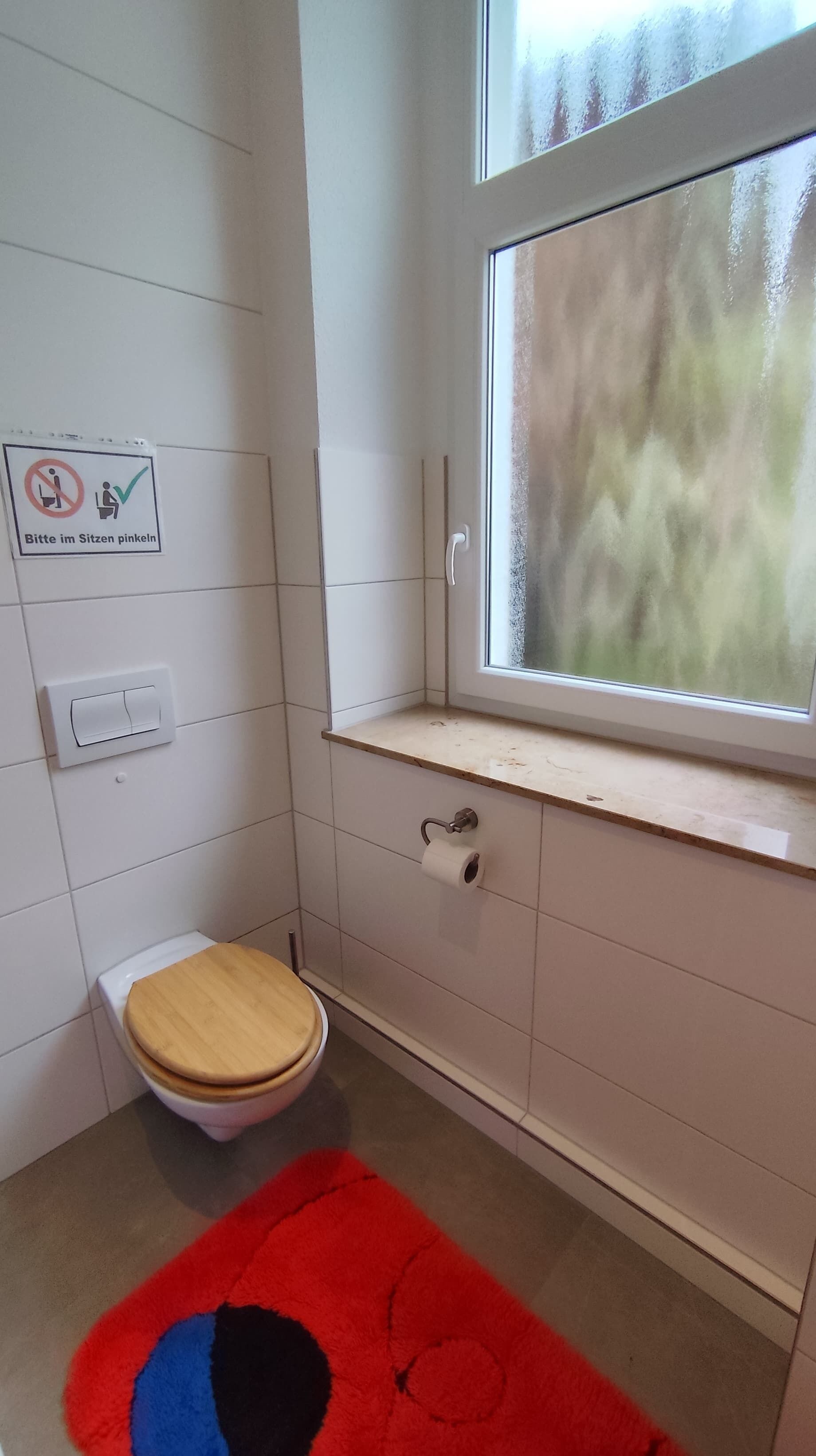A small bathroom with a toilet, a window, a towel rack, and a sign instructing to sit on the toilet seat.
