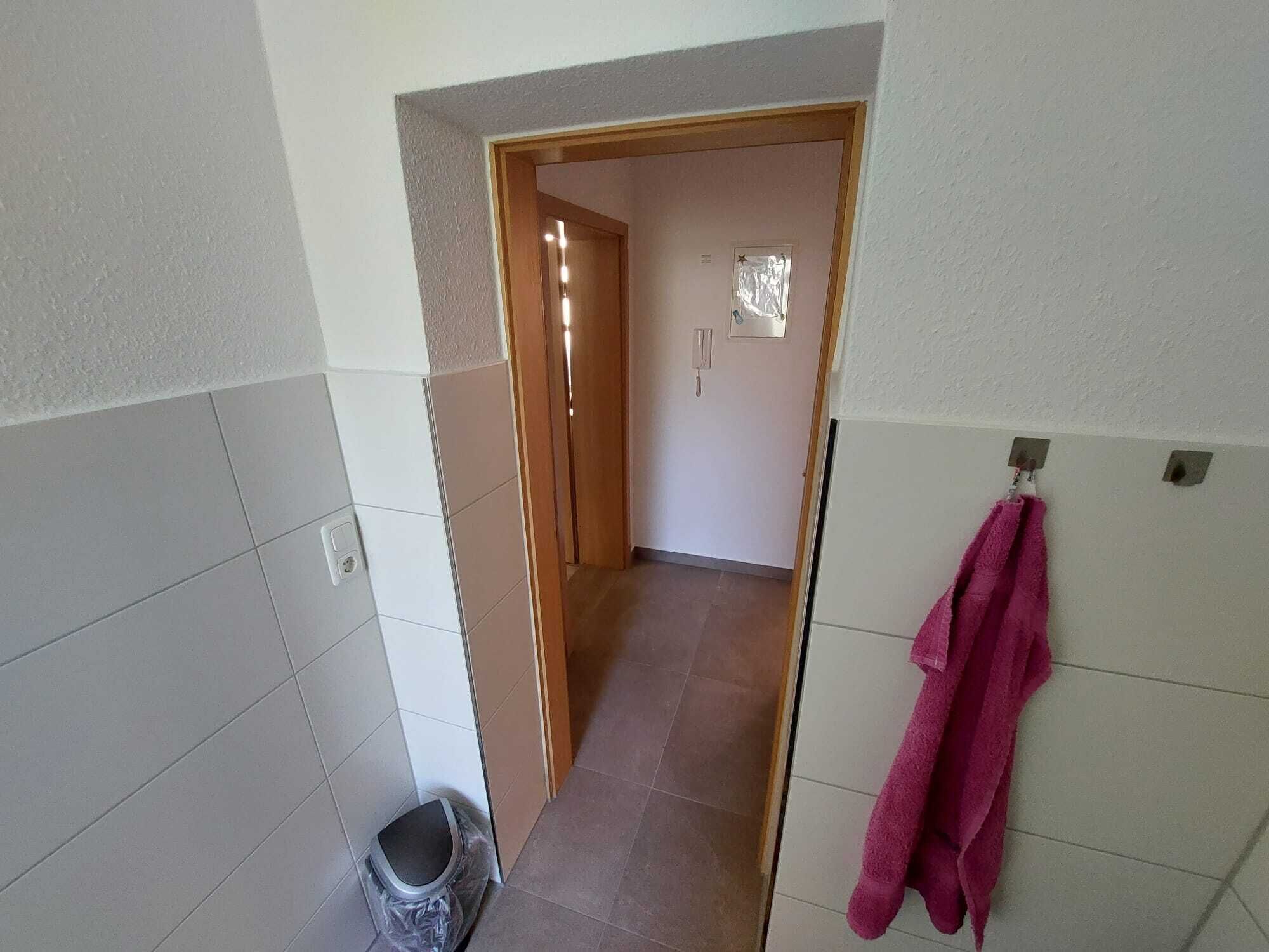 Un pasillo estrecho con una puerta de madera que da a una habitación. Una toalla rosa cuelga de un gancho en la pared derecha.