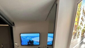 A hotel room with a bed, two rolled towels, a television on the wall, and a window with a view of a building. - MPods Málaga (Málaga)