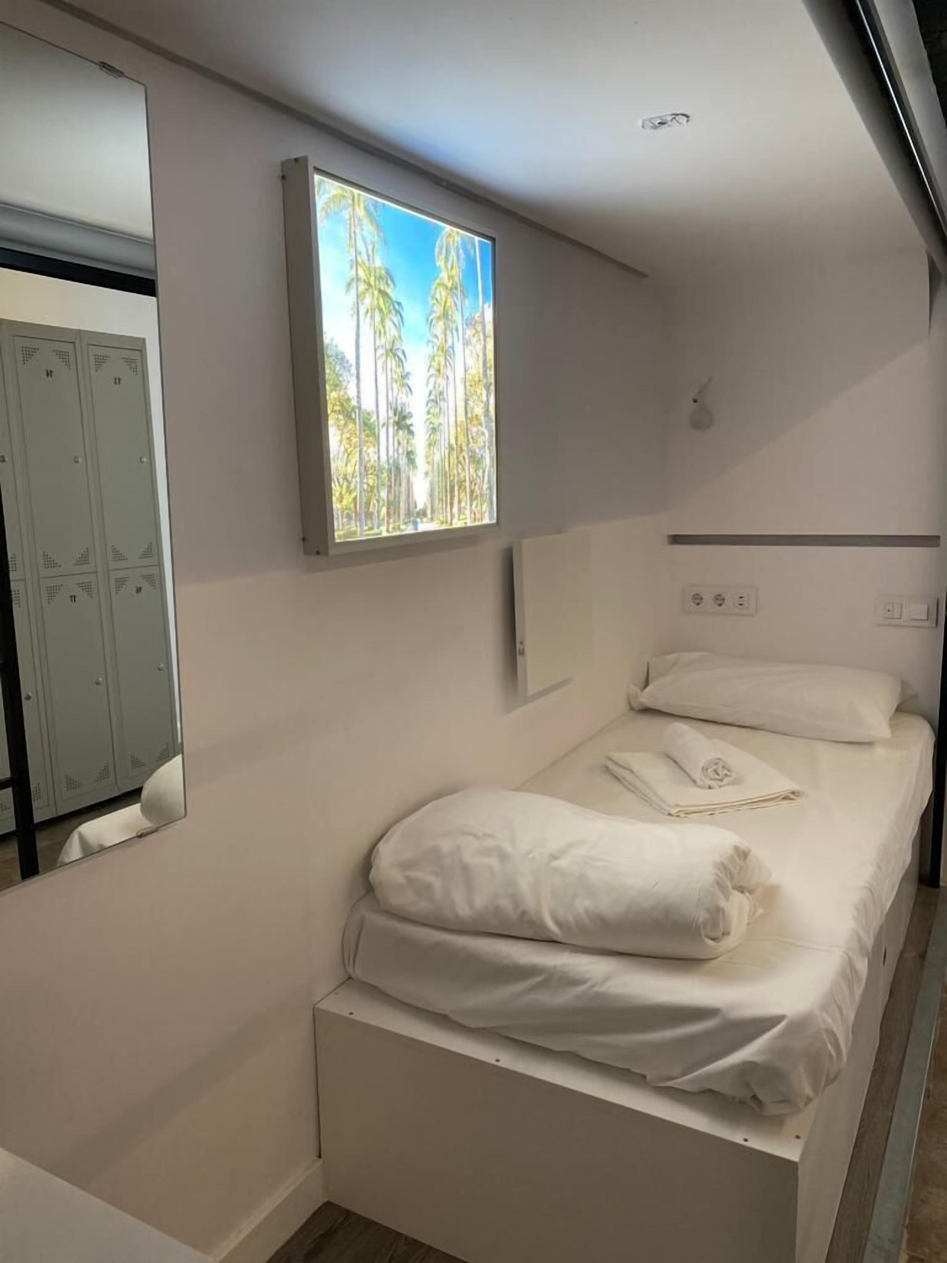 Une chambre avec un lit simple, une fenêtre donnant sur des palmiers et une armoire.