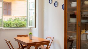 Un coin repas avec une table et des chaises en bois, une fenêtre donnant sur un bâtiment et de la verdure, et un meuble vitré contenant de la vaisselle.