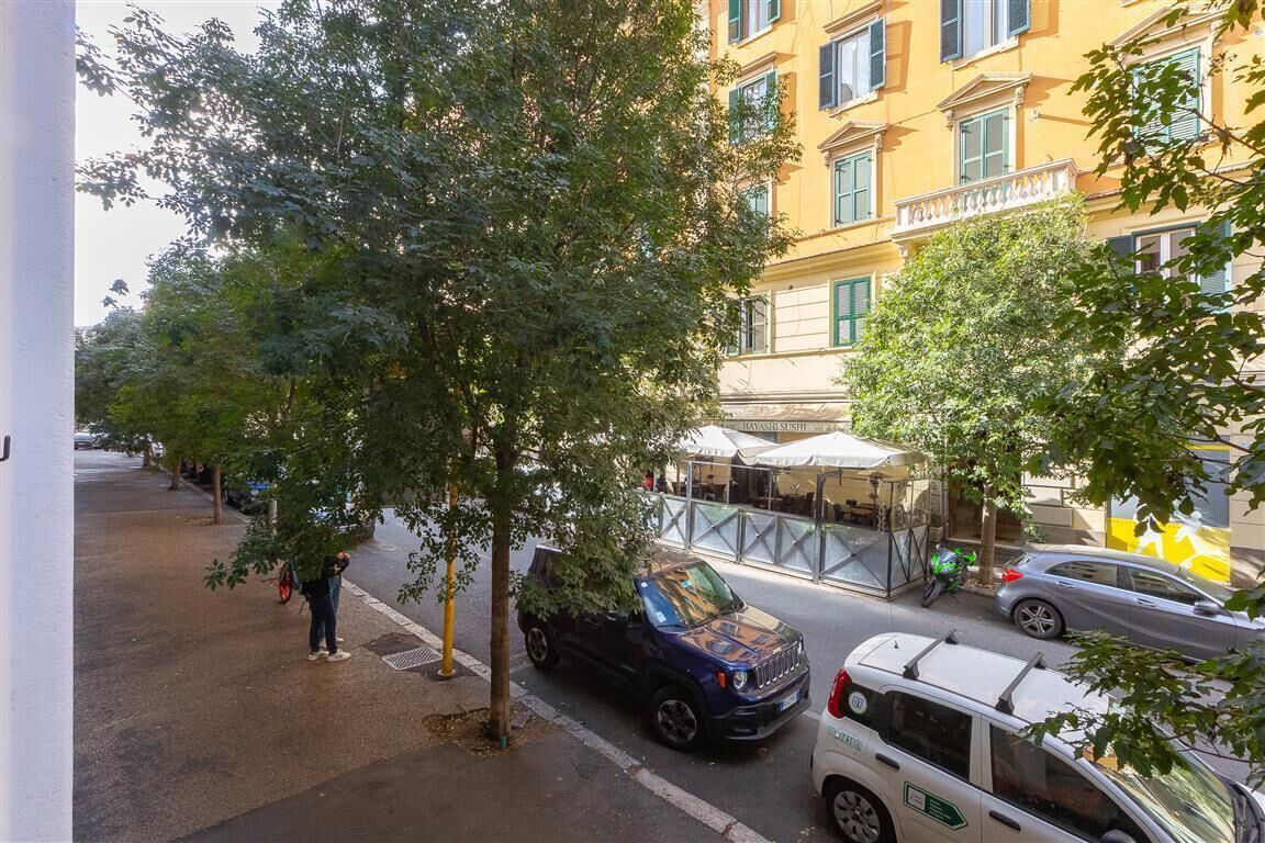 A street view with parked cars, a tree-lined sidewalk, and a building with a yellow facade.