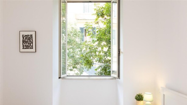 Une chambre à coucher avec un lit, une fenêtre donnant sur des arbres et des immeubles, une lampe de chevet et un tableau encadré au mur.