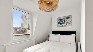 A bedroom with a bed, a window with a view of a building, a wall-mounted artwork, and a ceiling light fixture. - Contemporary Oasis in Paddington III (London)