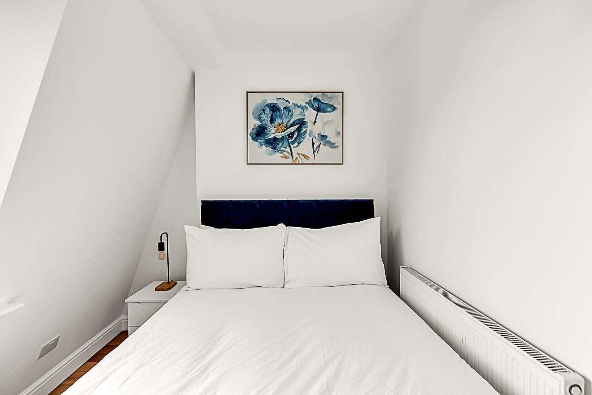 A bedroom with a white bed, a dark headboard, a nightstand with a lamp, and a framed artwork above the bed.
