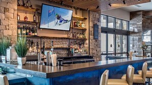Zona de bar con pared de piedra, estantes con botellas, una pantalla de televisión y taburetes.
