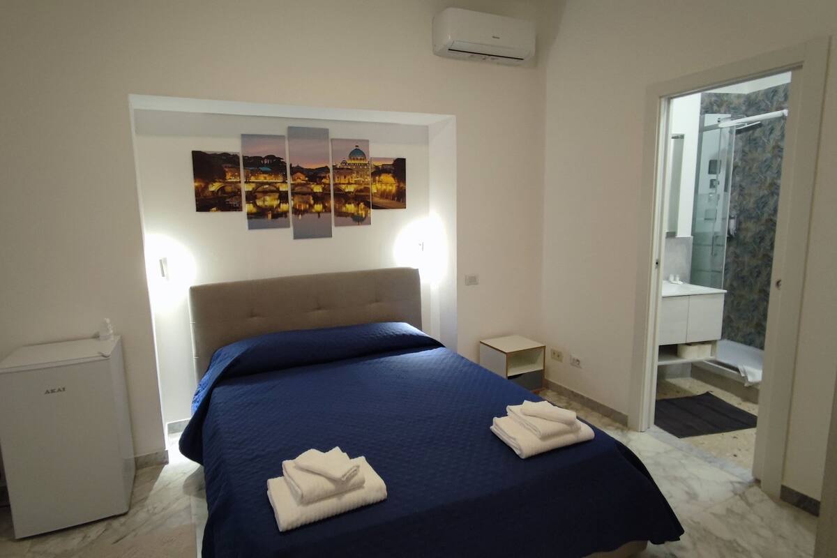 A bedroom with a bed, a small bedside table, a wall-mounted air conditioner, and a door leading to a bathroom.