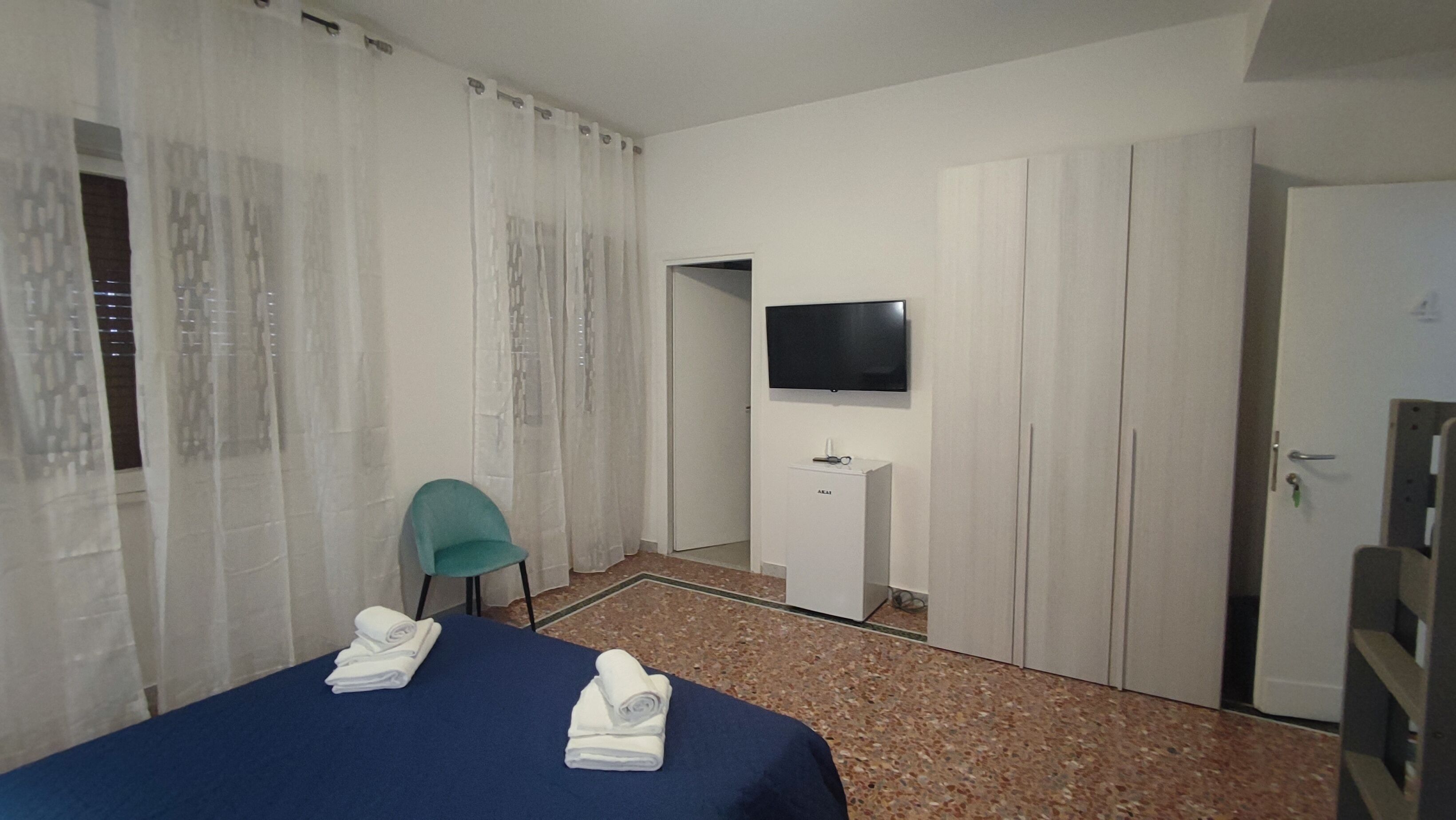 Una camera d'hotel con un letto, una sedia, una TV a muro, una scrivania e un armadio.