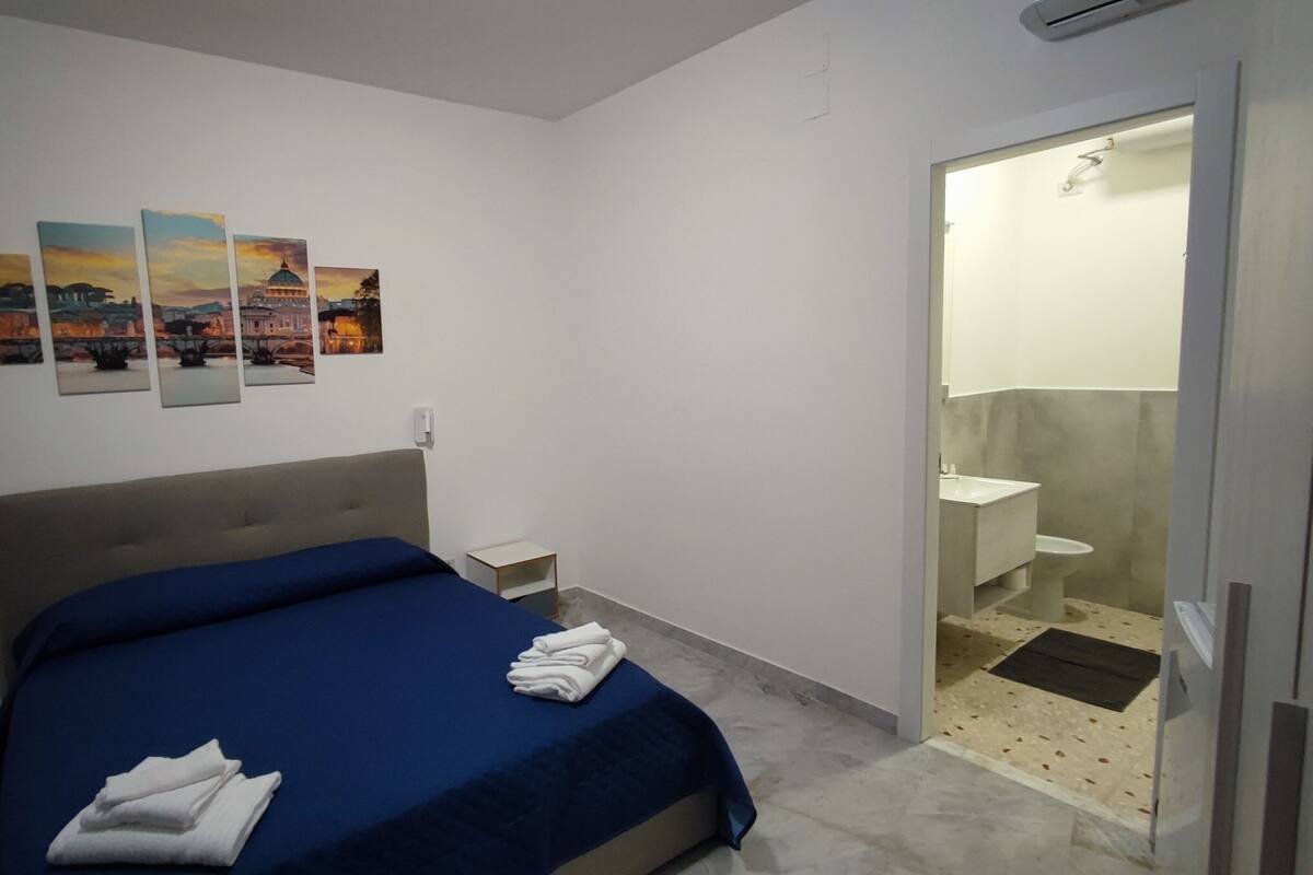 A bedroom with a bed, a nightstand, a small table, and a bathroom with a sink and toilet.