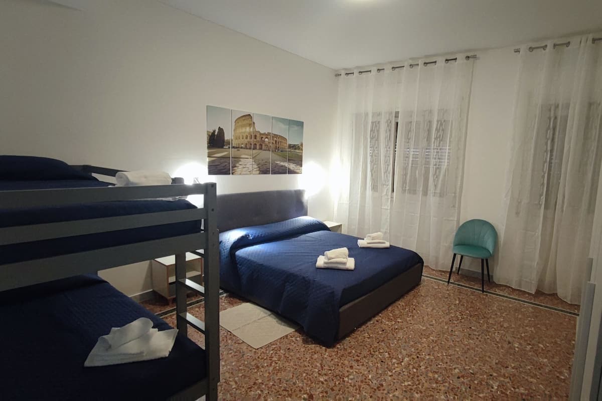 A room with bunk beds, a single bed, a chair, a window with curtains, and a picture of a historical building.