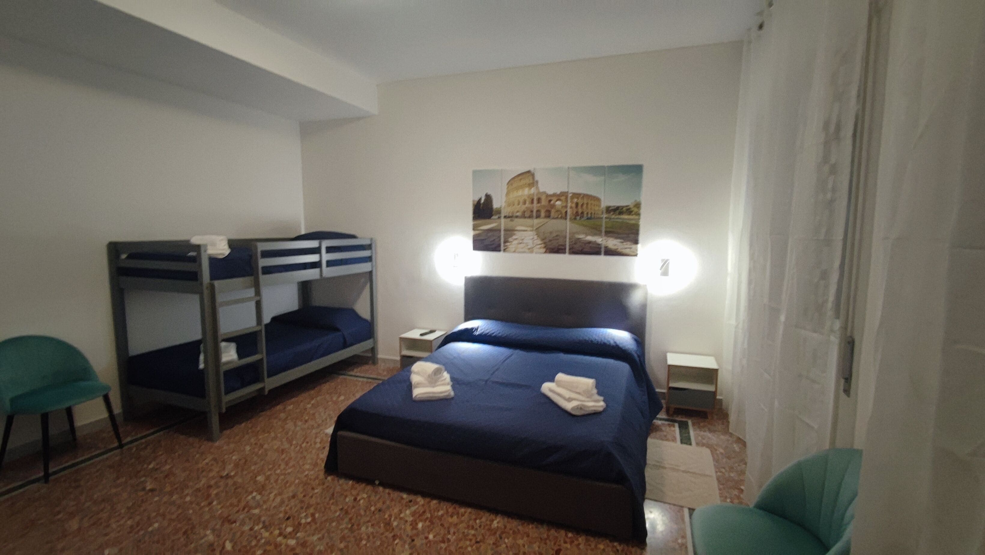 A room with bunk beds, a single bed, a painting of a historical building, and a small table with a lamp.