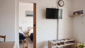 Ein modernes Hotelzimmer mit Bett, Nachttisch, Fernseher und einer kleinen Schreibtischzeile mit Stuhl und Blumenvase.