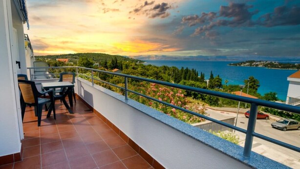Ein Balkon mit Blick auf einen Sonnenuntergang über dem Meer, ein Tisch mit Stühlen und ein Geländer.