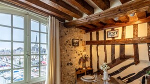 A room with a stone wall, wooden beams, a view through a window, and a table with a vase of flowers.