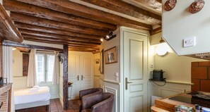 A rustic room with exposed wooden beams, a bed with white linens, a brown leather chair, a white door, and a kitchen area with a coffee machine.