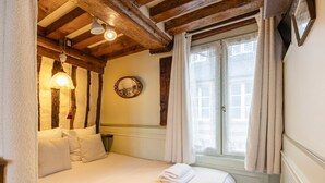 A cozy bedroom with a wooden ceiling, a bed with white linens, a round mirror, and a window with curtains.