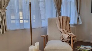 Una habitación con una silla cubierta con un paño color beige, una mesa con velas y un cuadro enmarcado, y una lámpara de pie.