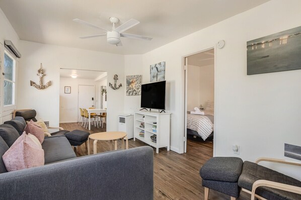 A modern living room with a grey sofa, a small round table, and a TV on a white stand. There is a dining area with chairs and a white cabinet with shelves.