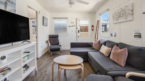 A modern living room with a flat-screen TV, a grey sofa with cushions, a wooden coffee table, and a ceiling fan.