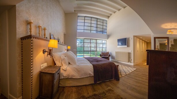 A spacious bedroom with a large bed, a wooden dresser, a TV, and a view of the outdoors through a large window. - Luxury Villa in Costa Brava with Private Pool, Tennis Court, and Sauna
 (Pantans de Montbarbat)