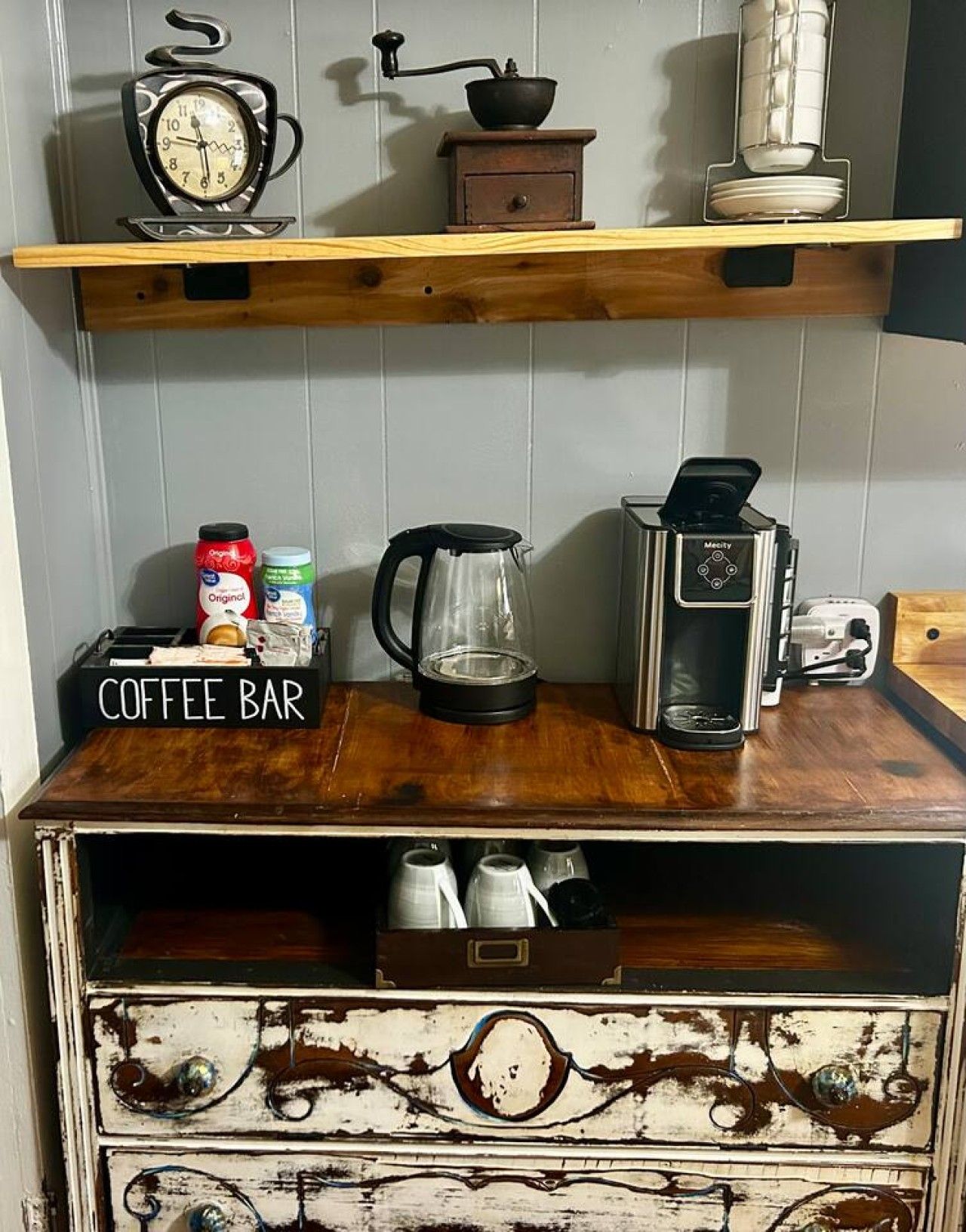 A coffee bar with a vintage clock, a coffee grinder, a kettle, and a coffee machine on a wooden shelf.
