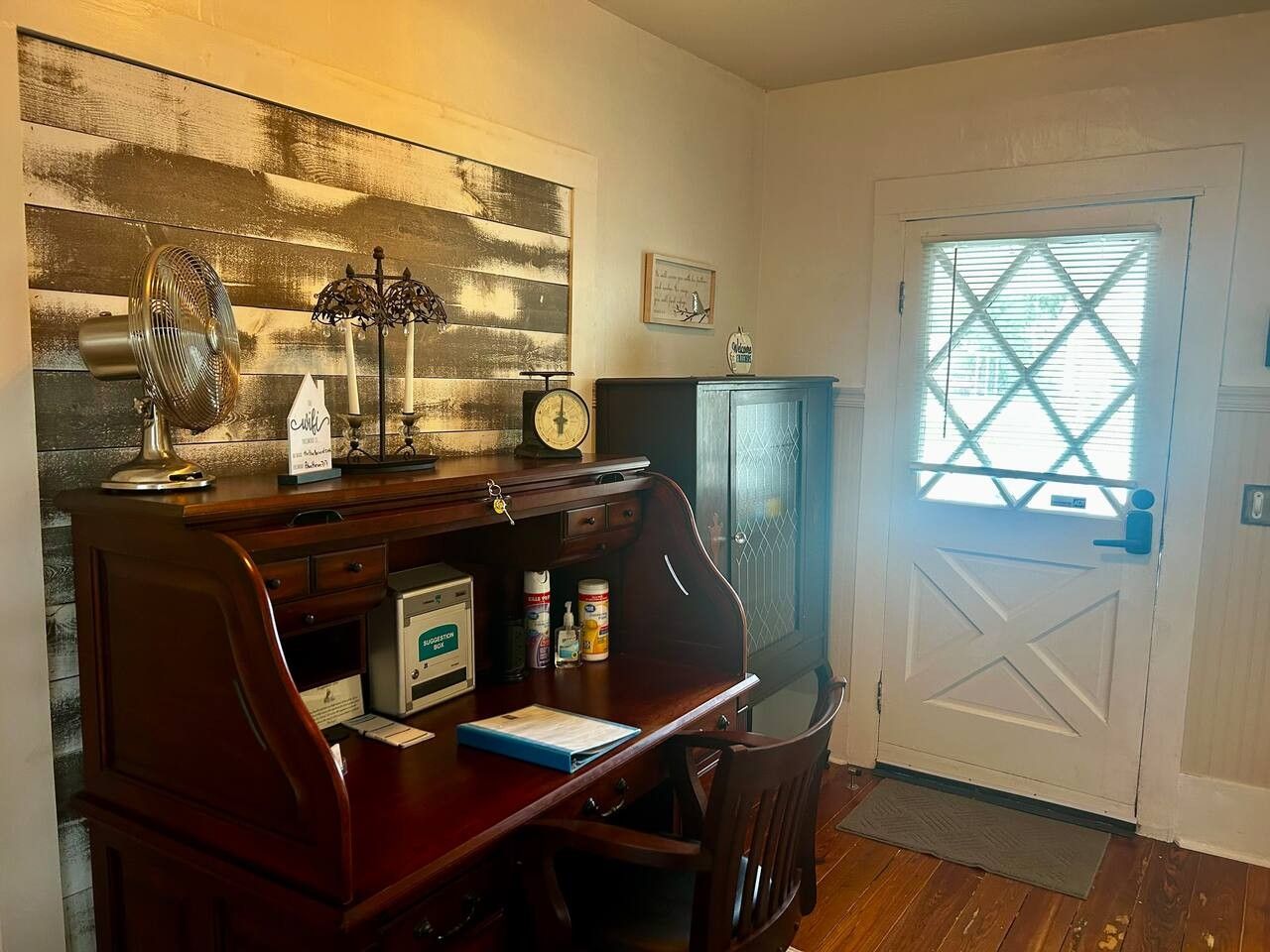 Een kamer met een houten bureau, een klok, een raam met jaloezieën en een deur met een kruisdecoratie.