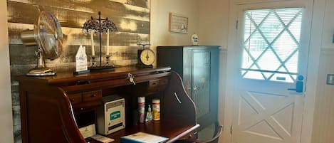 Une pièce avec un bureau en bois, une horloge, une fenêtre avec des stores et une porte ornée d’un motif en forme de croix.