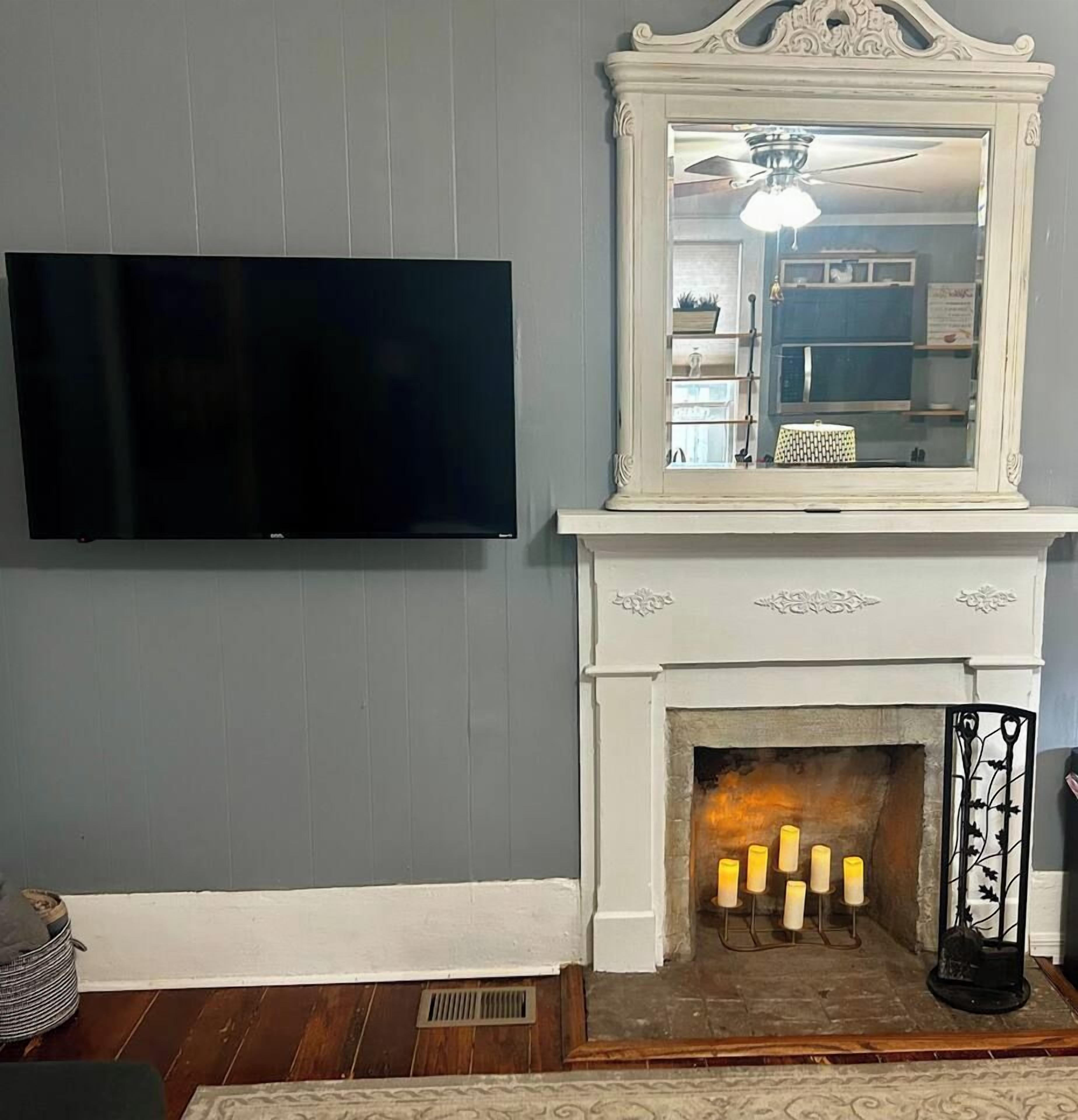 Een kamer met een wandgemonteerde flatscreen televisie, een witte haard met een spiegel erboven en een klein kandelaarhoudertje met kaarsen.
