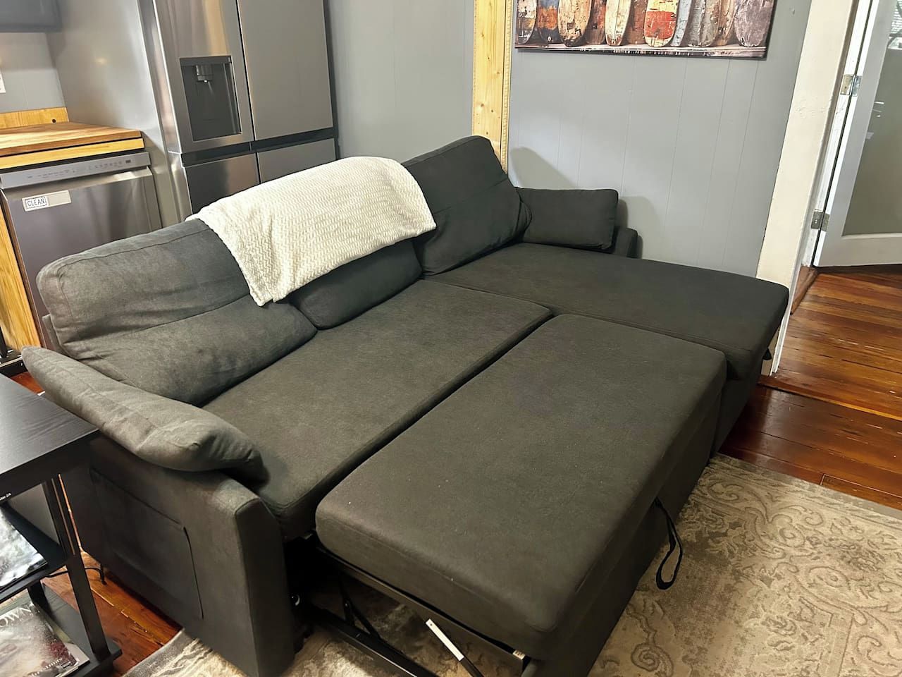 Ein graues Sofa mit einer weißen Tagesdecke, ein Holznachttisch und eine Küchenzeile mit Kühlschrank und Geschirrspüler.