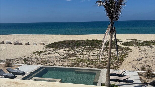A pool area with lounge chairs and a palm tree, overlooking a beach with a clear blue sky.