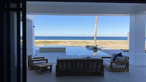 A modern interior with a large glass door leading to a beach view. Inside, there are wooden furniture pieces and a small pool area.