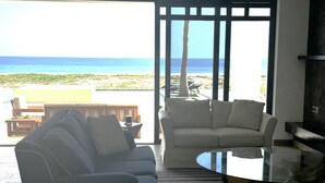 A living room with a sofa, armchair, and coffee table, overlooking a beach with a view of the ocean.