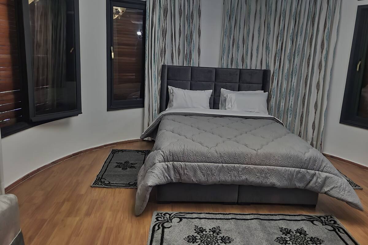 Un dormitorio con una cama grande, un cabecero y dos ventanas con persianas.