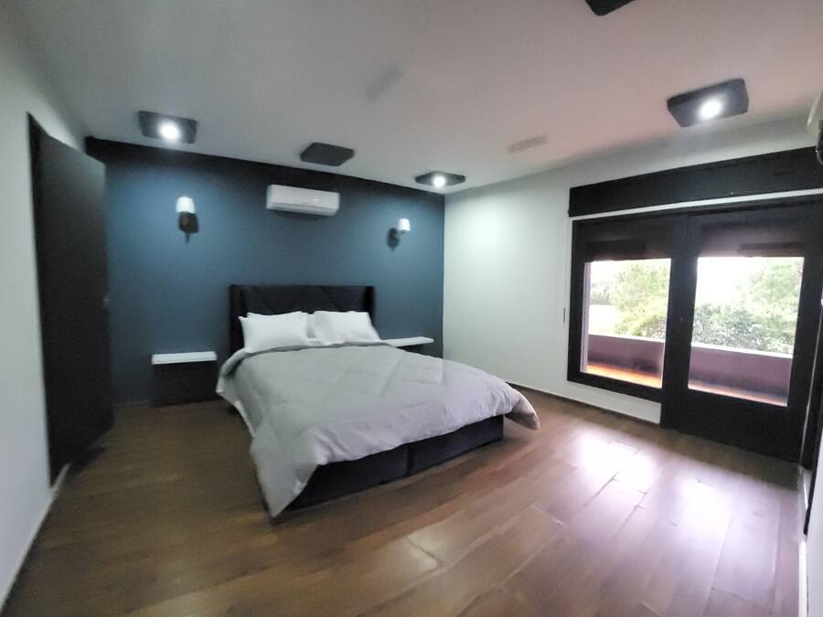 A bedroom with a bed, a nightstand, a wall-mounted TV, and a large window with a view of greenery.