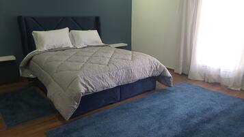 A bedroom with a bed, a nightstand, a wall-mounted lamp, and a window with curtains.