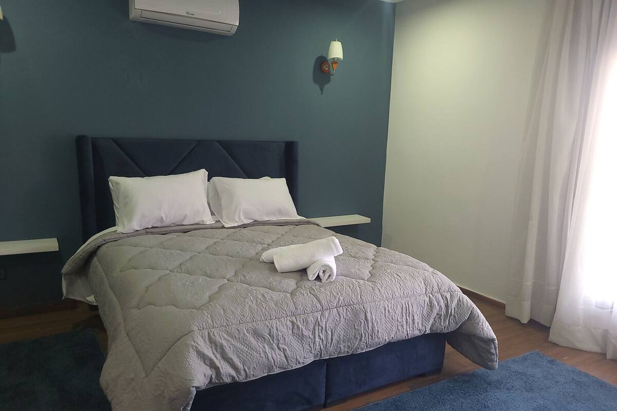 Una cama bien hecha con colcha gris, almohadas blancas y una toalla blanca enrollada.