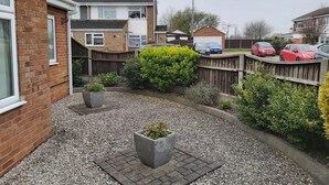 A residential backyard with a gravel pathway, two potted plants, a wooden fence, and several parked cars. - Luxurious home for long stay  (East Tilbury)