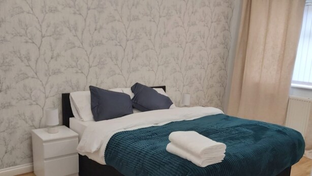 A neatly made bed with a teal blanket and white pillows, a bedside table with a lamp, and a wall with a tree pattern. - Luxurious home for long stay  (East Tilbury)