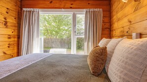 A wooden cabin room with a bed, a window with curtains, and a view of trees outside.
