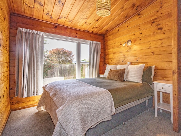 Quarto de cabana de madeira com cama, mesa de cabeceira e janela com cortinas.