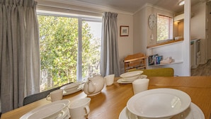 A dining area with a wooden table set for four, a window with curtains, a clock on the wall, and a view of greenery outside.