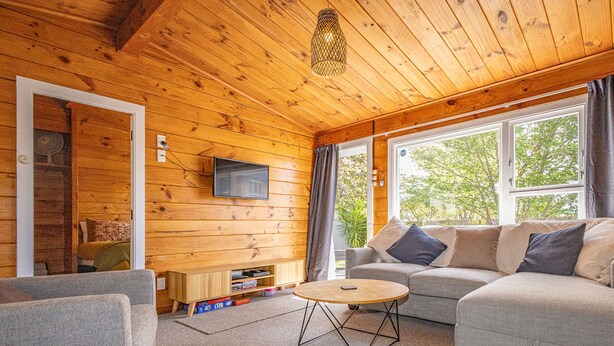 A cozy cabin interior with a wooden ceiling, a sofa, a small round table, and a TV mounted on the wall.