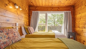 A wooden cabin bedroom with two beds, a window with curtains, and a small table.