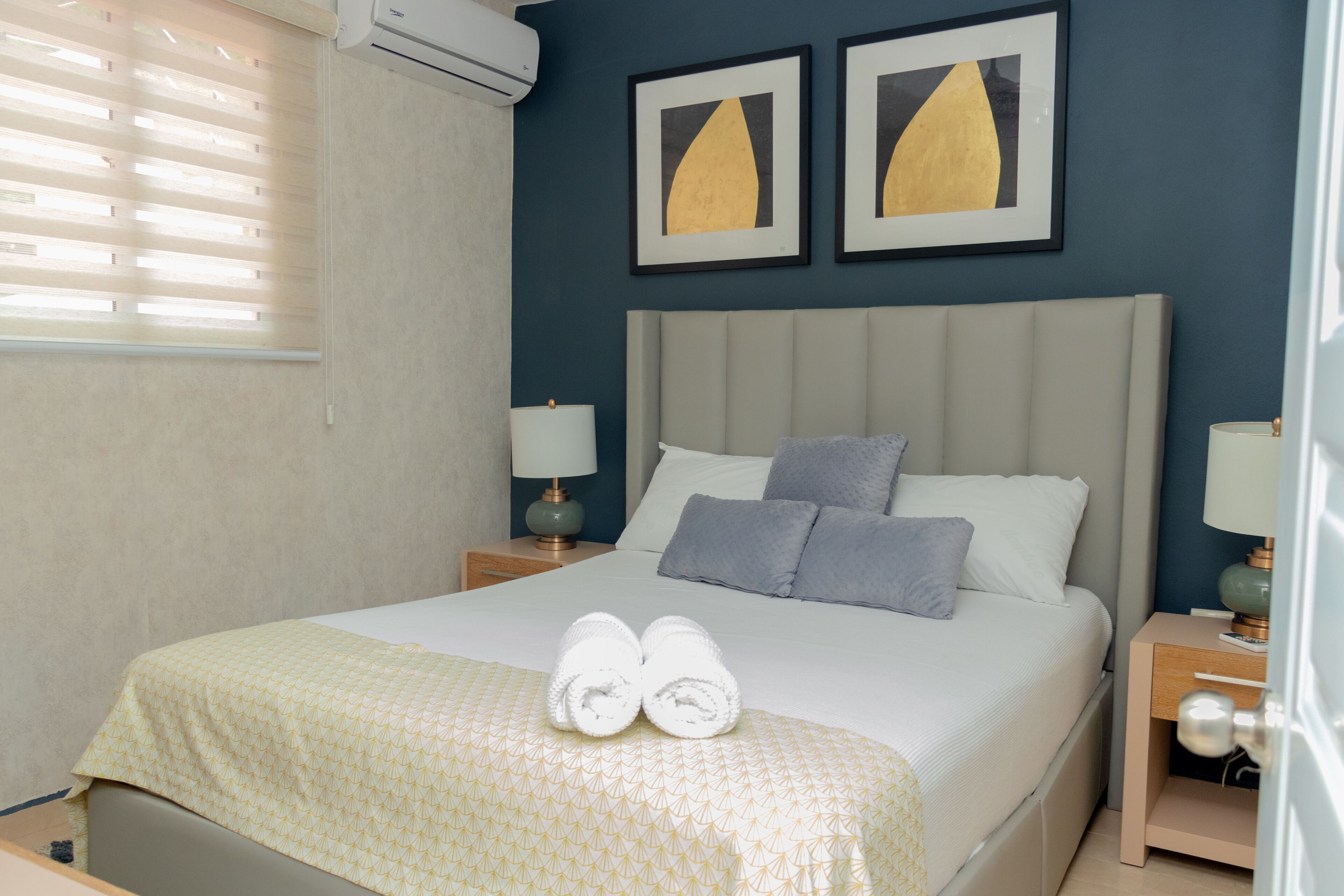 Cama bem arrumada com colcha estampada, dois travesseiros e um par de toalhas enroladas. Possui duas mesas de cabeceira com luminárias, e a parede de destaque é azul, com duas fotos emolduradas.